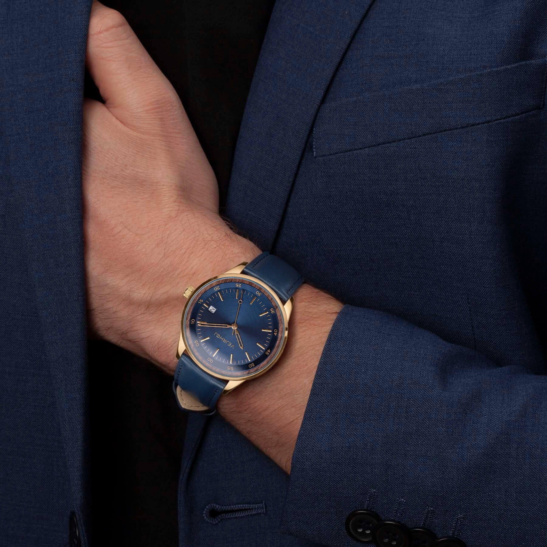 A close-up of a men's wristwatch with a blue dial and blue strap, worn on a person's wrist. The watch has a gold-tone case and is styled on a person wearing a dark blue suit jacket and a black shirt.