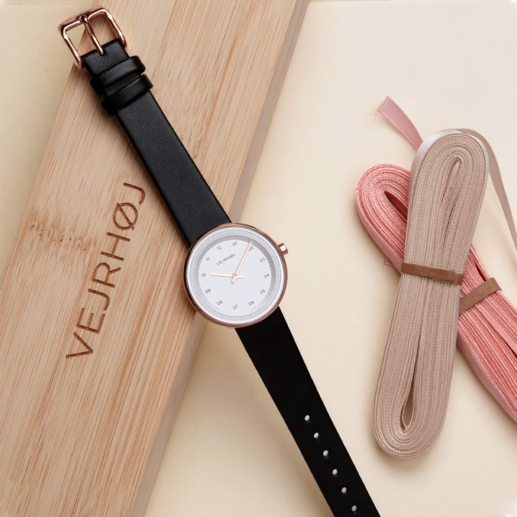 white ladies watch with a black strap and rose gold case on a wooden box with a VEJRHØJ  logo .