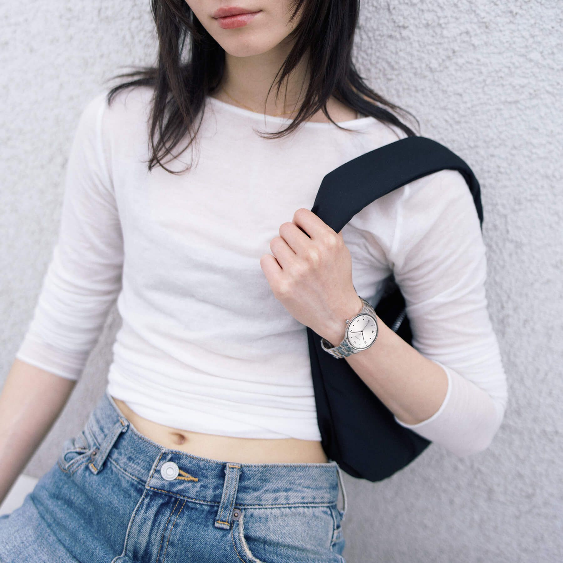 women wearing a VEJRHØJ steel watch and a white cropped top and blue jeans with a black strap over shoulder against a light gray background