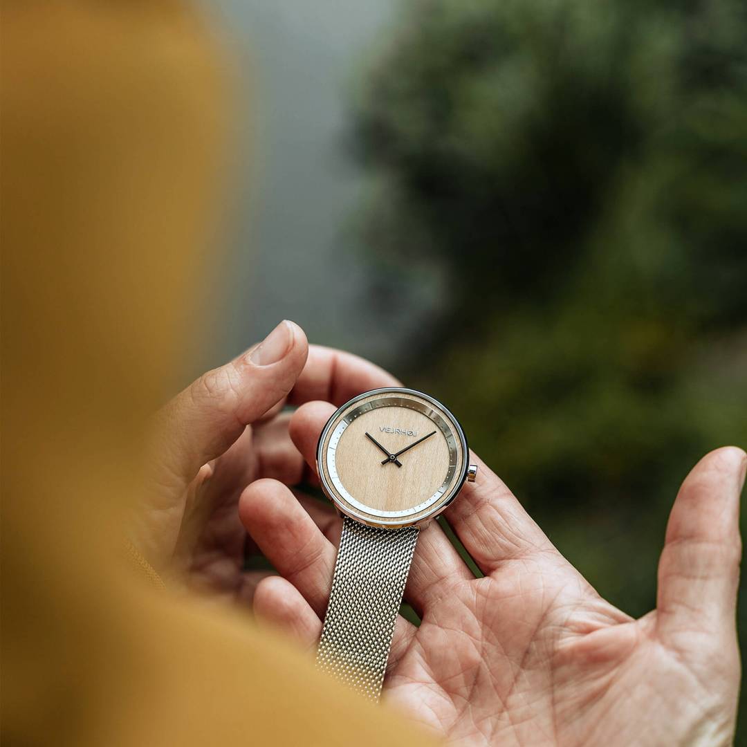 Person holding and looking at a simple wooden watch with silver casing and steel mesh band